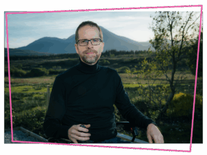 Matthew in his wheelchair wearing a black polar neck jumper in front of the mountains