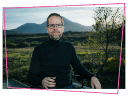 Matthew in his wheelchair wearing a black polar neck jumper in front of the mountains