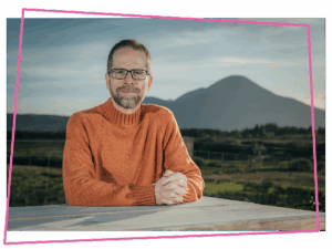 Matthew sat at a table in an orange jumper with mountains in the background