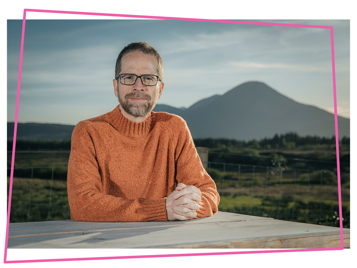 Matthew sat at a table in an orange jumper with mountains in the background